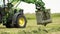 Green tractor picks up stack of grass with metal claw at farm field