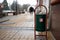 green trash bin with a red sticker near the entrance to the building on the background of a blurred park