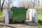 A green trash can stands on a site with a white concrete fence in a public park. Garbage collection and disposal using a plastic