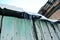 Green wooden old wall with dark shabby roof with icicles and white snow on top, view from ground on blue clear sky background