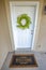 Green wreath hanging on the white front door of a home with a Welcome doormat