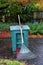 Green yard waste container, garden rake and shovel, sitting on a wet driveway, garden in background, fall cleanup