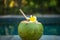 Green young coconut close up with bamboo straw and tropical flower frangipani on the edge of swimming pool