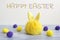 Greeting postcard with happy Easter. a yellow toy hare and round yellow and purple wool balls on gray wooden boards and on a gray