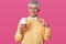 The grey haired senior in bright shirt and bowtie holds big cup of coffee. Elderly man stands with spectacles against studio wall