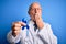 Grey haired senior doctor man holding colon cancer awareness blue ribbon over blue background cover mouth with hand shocked with