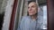 Grey haired senior man stands behind balcony glass and looks with sorrow.