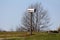 Grey metal mailbox mounted on wooden pole on top of small hill surrounded with grass and large tree on clear blue sky background