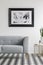 Grey settee on checkered floor in white living room interior with gold table and poster. Real photo