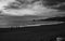 Greyscale of a beach surrounded by the sea and rocks under a cloudy sky in the evening