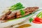 Grilled Cevapcici is a Balkan national dish. Close the row of fried beef kebabs on a white rectangular dish with vegetables