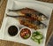 Grilled Torpedo Scad fish Ikan Cencaru Bakar in Malay with hot soy sauce, hot tamarind juice and fresh cucumber on white plate