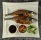 Grilled Torpedo Scad fish Ikan Cencaru Bakar in Malay with hot soy sauce, hot tamarind juice and fresh cucumber on white plate