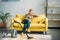 grimacing woman sitting on sofa and looking at camera vacuum cleaner on floor