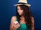 Grimacing young woman in hat looking on mobile phone with question sing on the face on blue background