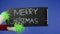 The Grinch's green haired hand holds out a black sign that reads Merry Christmas on an isolated blue background