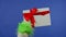 The Grinch's green haired hand holds out a white gift box with a red bow on an isolated blue background. Gift