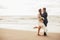 Groom in a chic suit and a beautiful bride in a wedding gown walking along the beach. Newlyweds kisses in protective medical masks