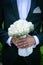 The groom in a gray suit,white shirt with a ring on his finger holding a wedding bouquet of white roses in his hands in front of h