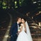 Groom reaches to the bride cheek while she stands with closed eyes in the park