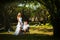Groom sits on a lawn on a chair in the middle of tropical trees, and the bride stands next to him