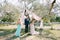 Groom stands with bride near a tree in a green grove in front of the wedding ceremony master