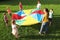 Group of children and teachers playing with rainbow playground parachute on green grass. Summer camp activity