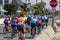 a group of cyclist riding along a street wearing colorful clothes with homes and cars and trucks along the street at Horny Corner