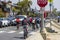 a group of cyclist riding along a street wearing colorful clothes with homes and cars and trucks along the street at Horny Corner