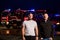 Group of firefighters, dressed in civilian clothing, stand in front of fire trucks during the night, showcasing a moment