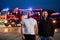 Group of firefighters, dressed in civilian clothing, stand in front of fire trucks during the night, showcasing a moment