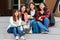 Group of four college student girls holding books, sitting and sharing time in front of outside school building with happy and fun