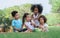 Group of four mixed race African and caucasian little cute kids sitting, playing in outdoor green park for picnic, eating fruit,