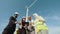 Group of four multiracial coworkers in safety helmets smiling on camera while staning on farm with huge windmills