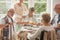 Group of friends with helpful carer sitting together at the table at nursing home dining room and eating cake