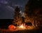 Group of friends warm their hands around campfire in woods near tent under an unusually starry sky