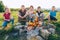 Group of Kids - Boys and girls cheerfully smiling and roasting sausages on long sticks over a campfire flame. Outdoor active time