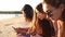 Group of millenial girls using smartphones laying together on beach towel near sea on summer sunset. Young women