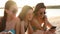 Group of millenial girls using smartphones sitting together on beach towel near sea on summer sunset. Young women