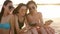 Group of millenial girls using smartphones sitting together on beach towel near sea on summer sunset. Young women