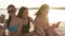 Group of millenial girls using smartphones sitting together on beach towel near sea on summer sunset. Young women