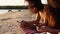 Group of millenial women using smartphones laying together on beach towel near sea on summer sunset. Young people