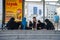 A group of Muslim women in black hijabs sit on the ground at a bus stop and enjoy their food