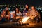 Group of people having fun sitting near bonfire outdoors at night playing guitar, singing songs and talking happily together