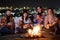 Group of people having fun sitting near bonfire outdoors at night playing guitar, singing songs and talking happily together