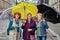 A group of three mature European women stand under umbrellas on a city street in rainy weather.