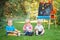 Group of three white Caucasian toddler children kids boys and girl sitting outside in summer autumn park by drawing easel