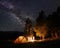 Group of tourists in night forest at tent camp look on burning fire under the starry sky