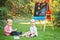 Group of two white Caucasian toddler children kids boy and girl sitting outside in summer autumn park by drawing easel