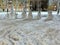 A group of various snow covered snowmen. With snowdrifts in front, trampled by various traces. On a city street.